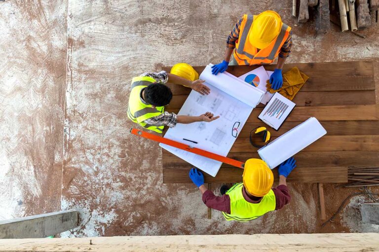 A person examines house plans at a construction site with framing materials and blueprints spread out on a table.